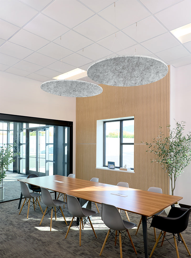 A modern conference room with a long wooden table, gray and black chairs, two large oval gray felt pendant lights hanging from the ceiling, a slatted wood accent wall, and large windows with a glass exit door.