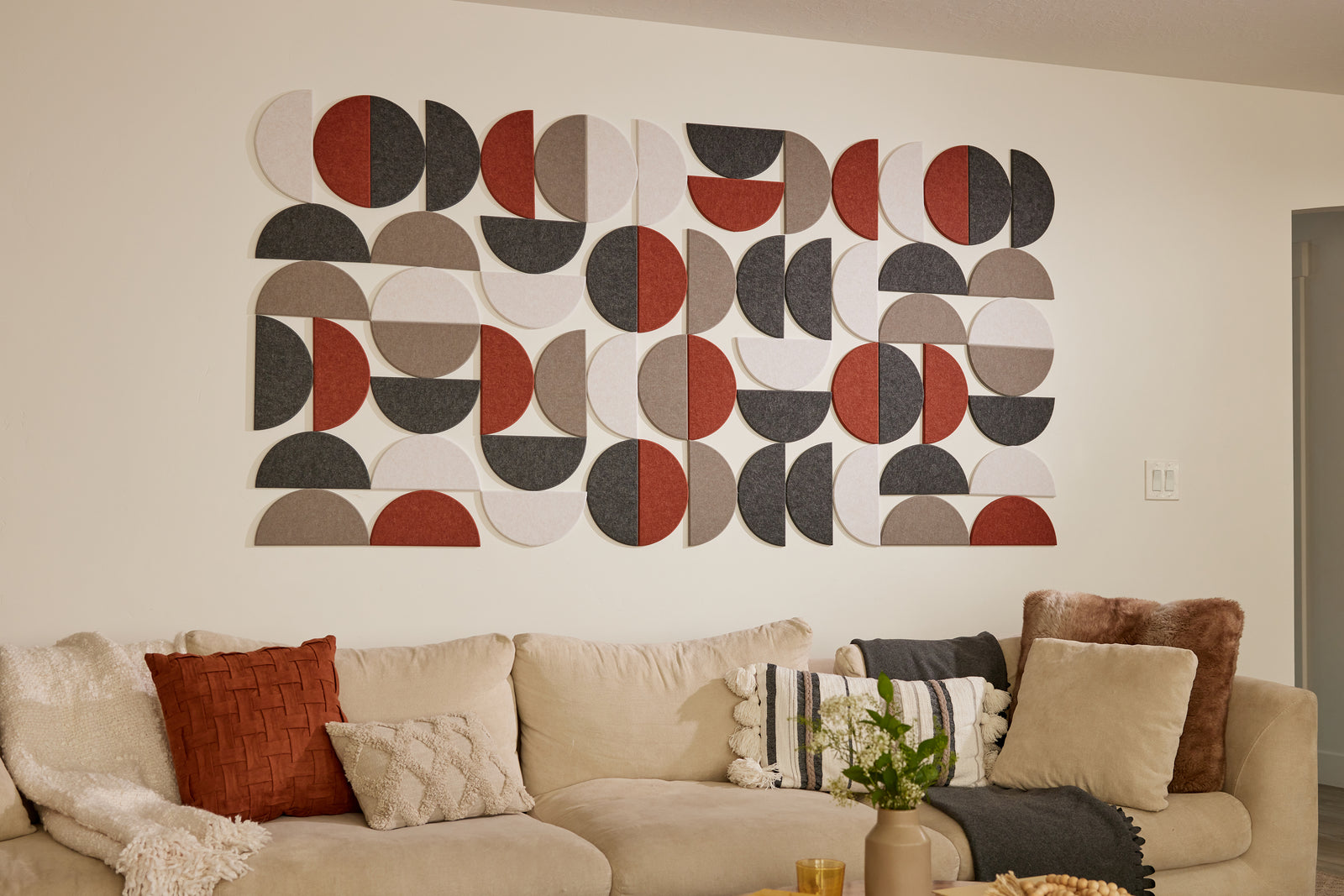 A Felt Right wall art design using half circles in shades of cream, grey, brown, and red on a wall above a couch with decorative pillows and blankets.