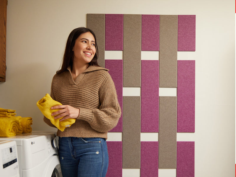 A Felt Right wall art design in shades of brown and pink on a white wall in a laundry room with a washer and a dryer and a woman doing laundry