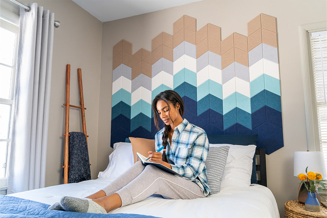 A Felt Right Wall art design resembling an ocean shoreline in shades of blue and tan behind a bed like a head board with a woman journaling on the bed.