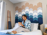 A Felt Right Wall art design resembling an ocean shoreline in shades of blue and tan behind a bed like a head board with a woman journaling on the bed.