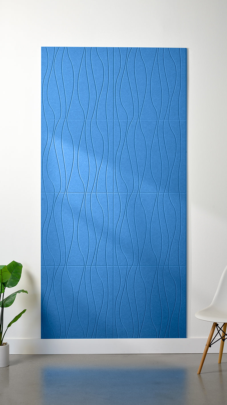 Bright blue Felt Right 4’ x 8’ PET felt panel with flowing lines engraved into the material against a white wall. A plant and chair are in the foreground.