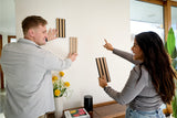 Two people holding and comparing wood slat samples with varying stain colors and finishes indoors.