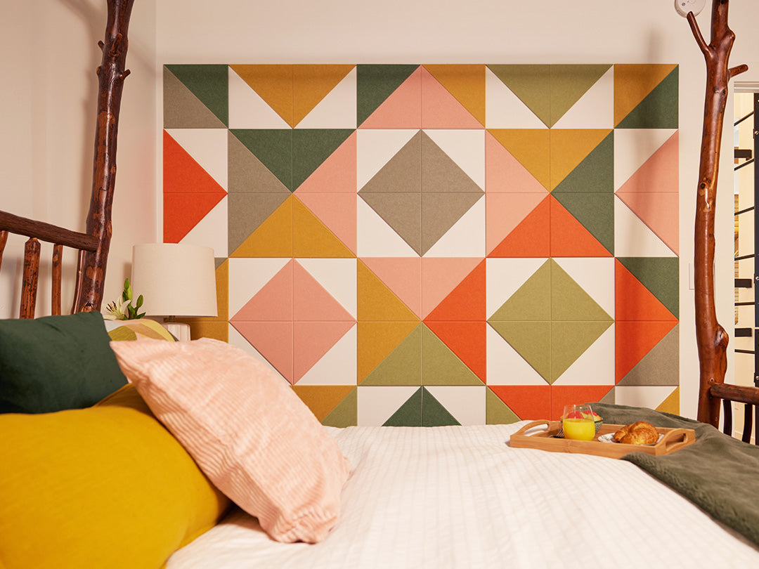 Felt Right wall art in shades of pink, green, orange, and yellow felt triangle felt tiles creating a pattern on a white wall with negative space covering a wall next to a bed in a bedroom