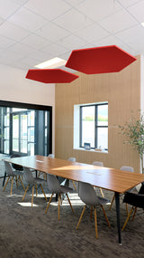 Two red hexagonal acoustic baffles suspended from ceiling in modern office conference room above wooden table.
