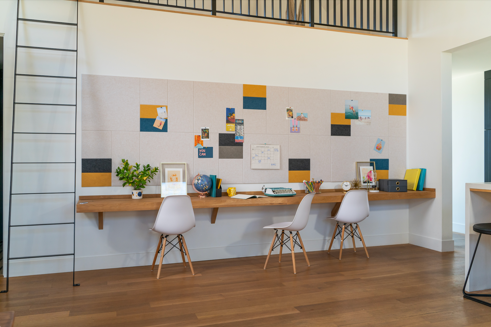 A Felt Right wall art design in shades of cream, yellow, blue, and black felt tiles on a white wall above a desk with three chairs.