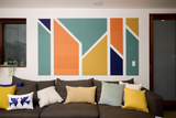 A Felt Right Wall Design on wall art with geometric shapes in orange, blue, green, and yellow mounted above a sofa with various colored cushions.