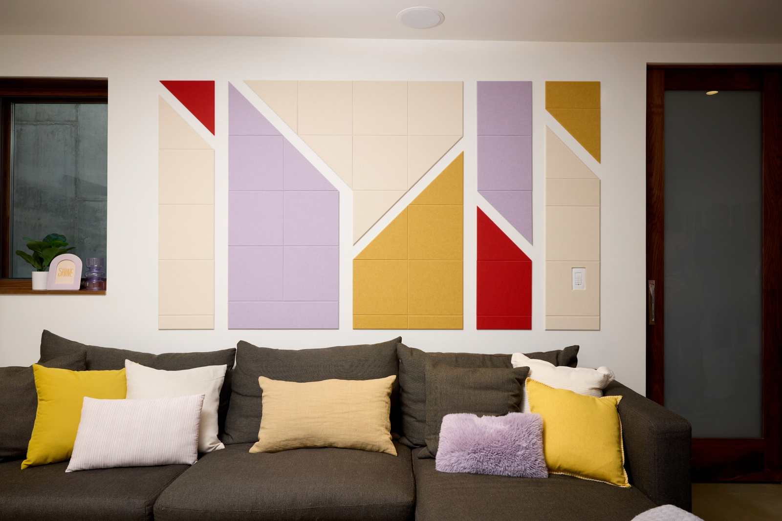 A Felt Right Wall Design colorful geometric patterned artwork hanging on a wall above a grey sofa, with cushions on the sofa, and a dimly lit ceiling light.