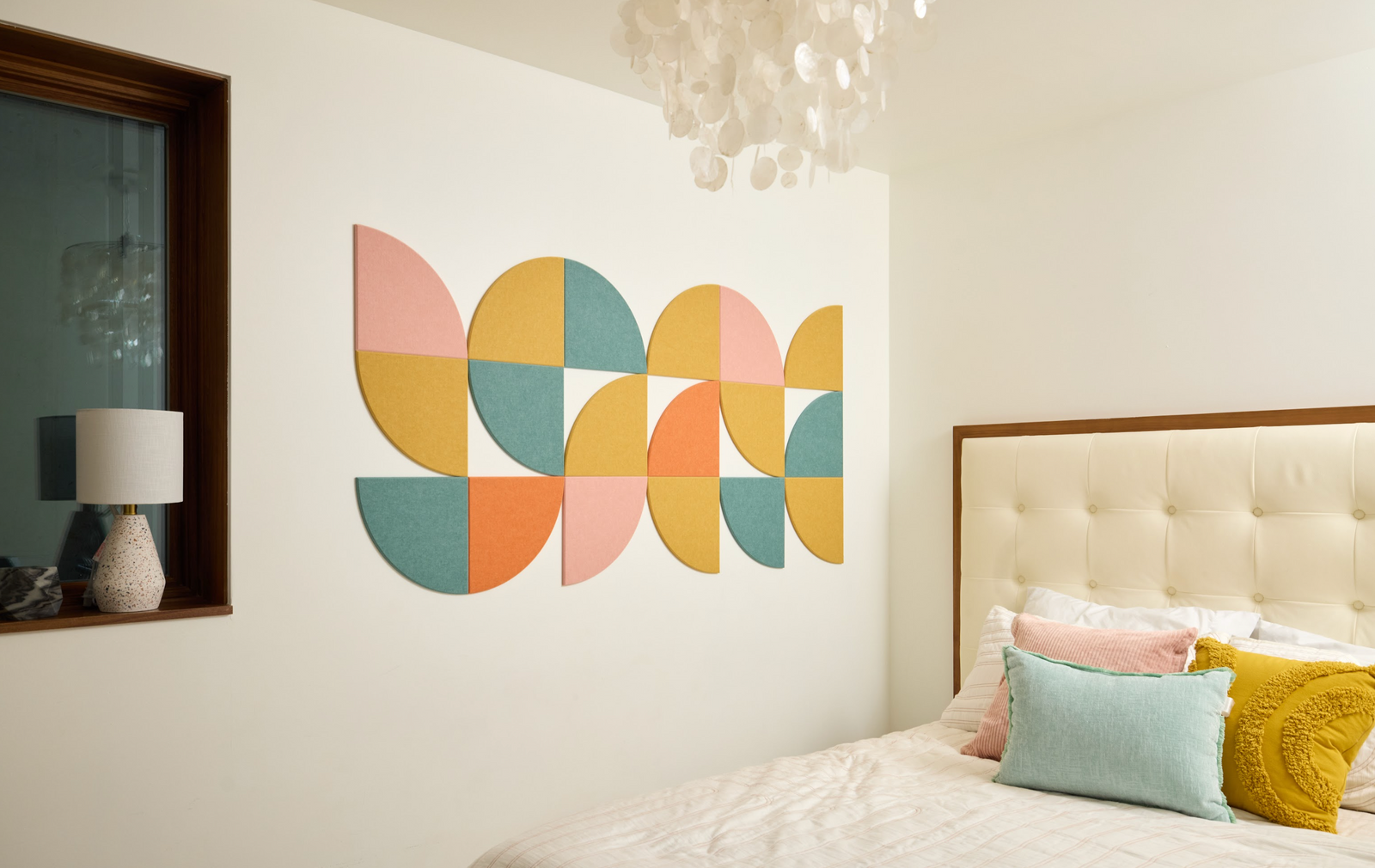 A Felt Right Wall Design quarter circle pieces in shades of pink, yellow, orange, and blue on a white wall in a bedroom next to a window with a lamp and a bed.