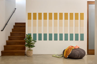 A Felt Right Wall Art design in shades of cream, yellow, and green next to stairs and a house plant.