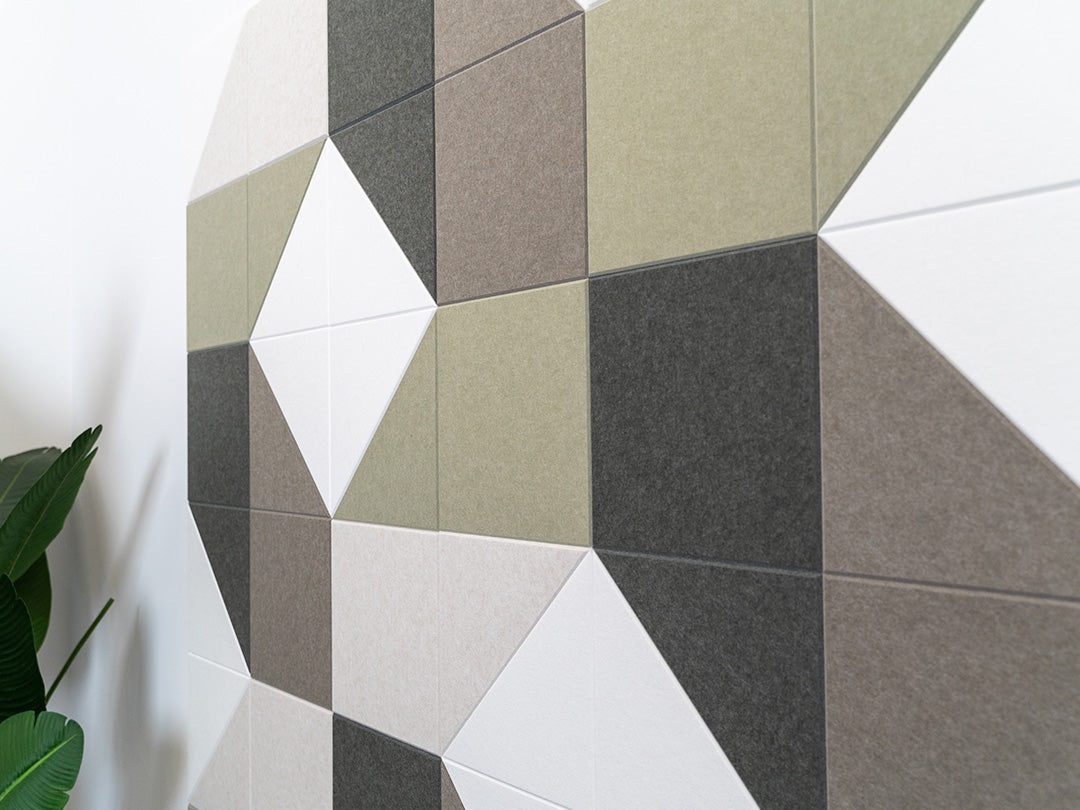 A Felt Right wall art design using shades of green, grey, and cream square and triangle tiles on a white wall next to a house plant.