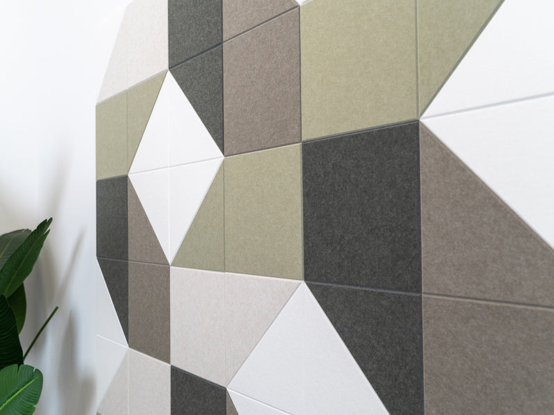 A Felt Right wall art design using shades of green, grey, and cream square and triangle tiles on a white wall next to a house plant.