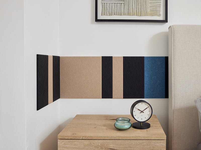 A Felt Right wall design in shades of black, cream, and blue felt tiles in a strip pattern going along a wall behind a bed and side table with a clock on it