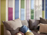 Felt right wall art design in shades of blue, green, purple, and cream felt tiles in a geometric shape lining the wall and the ceiling in a living room with a couch and some yarn.