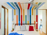 Felt right wall art design in a bedroom using shades of red, orange, blue, and cream felt tiles on a white wall and ceiling behind a bed with two light fixtures.