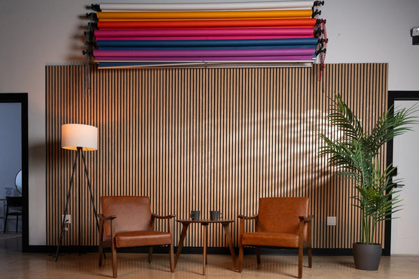 Two brown leather chairs with a small table between them against a wooden paneled wall with colorful fabric rolls above.