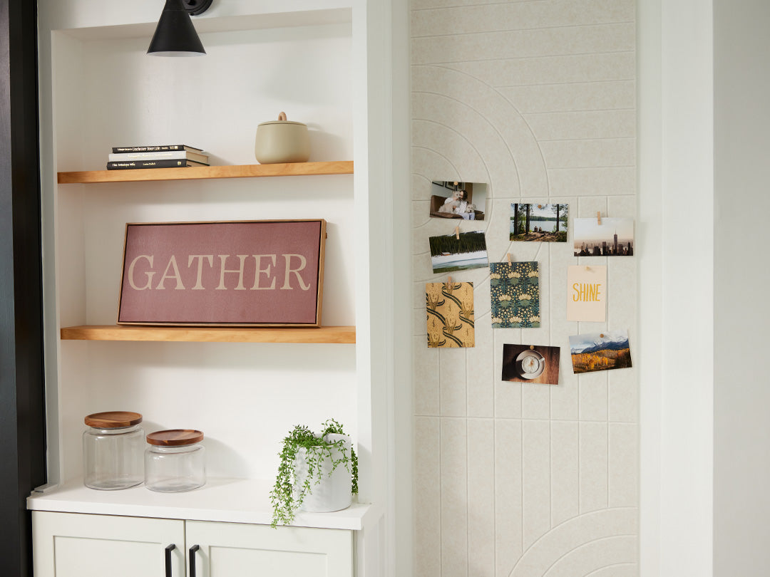 A Felt Right wall art design that has pictures pinned on the felt tiles that are in a shade of white or cream on a wall next to some shelves in a living room.