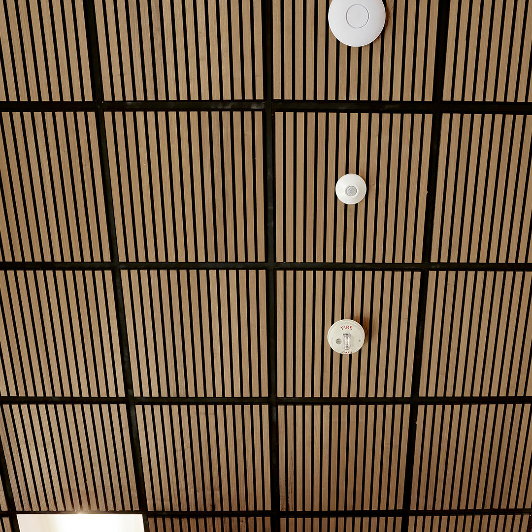 A close-up view of a slatted wood ceiling panel with a black grid frame and three white round smoke detectors attached vertically.