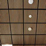 A close-up view of a slatted wood ceiling panel with a black grid frame and three white round smoke detectors attached vertically.