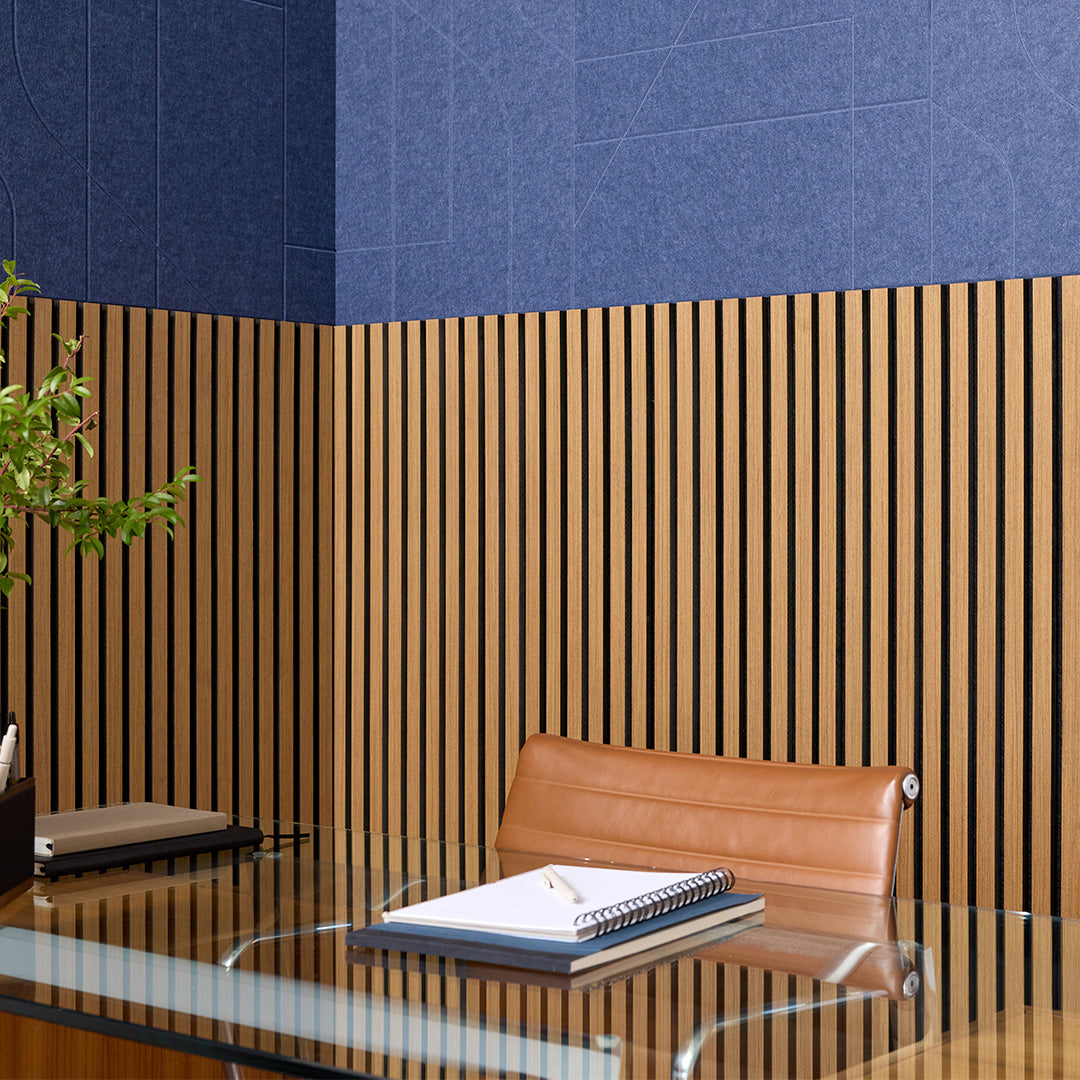 Modern office interior featuring a wall with a dark blue upper section and vertical wood slats on the lower section, with a glass desk, brown leather chair, and stacked notebooks in the foreground.