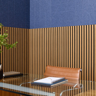 Modern office interior featuring a wall with a dark blue upper section and vertical wood slats on the lower section, with a glass desk, brown leather chair, and stacked notebooks in the foreground.
