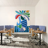 A modern office interior with two rows of desks and ergonomic chairs facing a large colorful abstract wall art piece on a white wall.