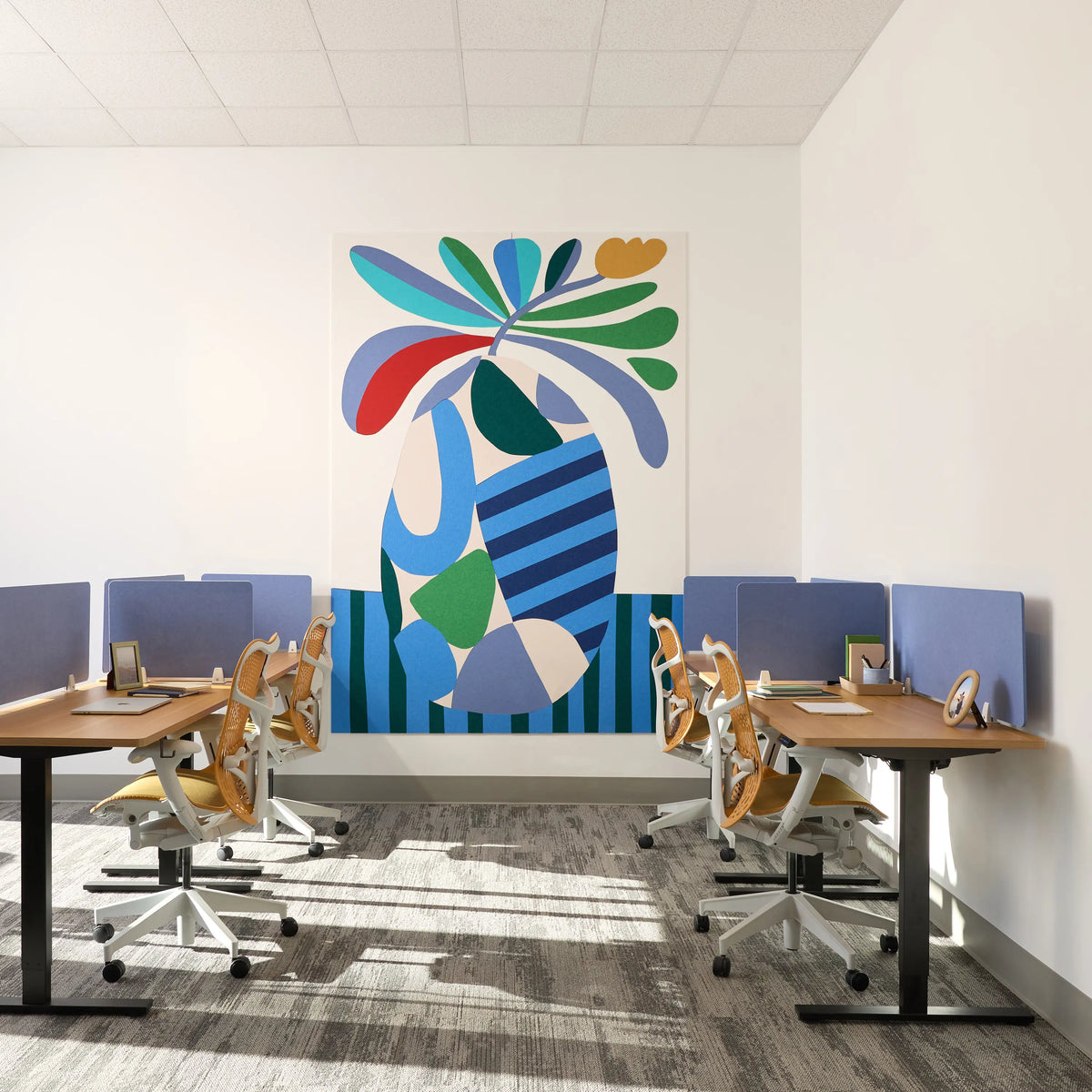A modern office interior with two rows of desks and ergonomic chairs facing a large colorful abstract wall art piece on a white wall.