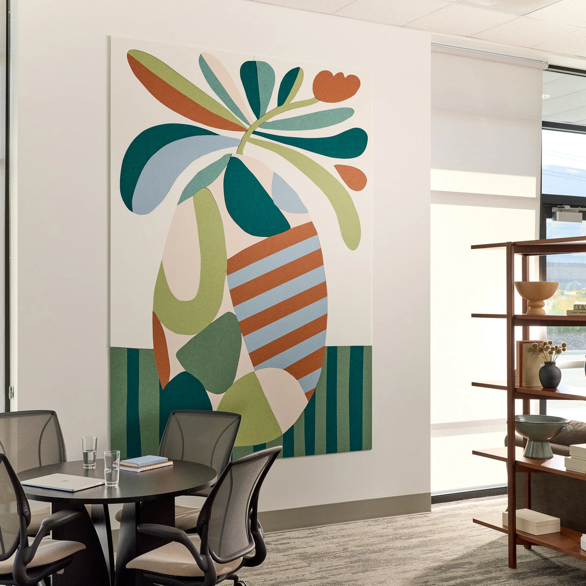 A large abstract wall art print featuring a stylized tropical leaf motif in teal, green, cream, orange, and light blue, displayed on a white wall in a modern office interior with a meeting table, chairs, and shelving visible.