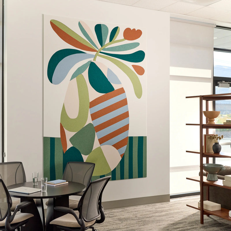 A large abstract wall art print featuring a stylized tropical leaf motif in teal, green, cream, orange, and light blue, displayed on a white wall in a modern office interior with a meeting table, chairs, and shelving visible.