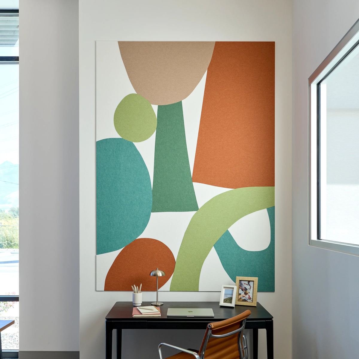 Large vertical acoustic wall art featuring an abstract geometric design with overlapping organic shapes in terracotta, sage green, teal, and beige on a white background, displayed on a wall above a black desk in a modern office setting.