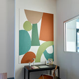 A large abstract wall art print with organic shapes in terracotta, sage green, teal, and beige on a white background, displayed above a black desk in a bright office space.