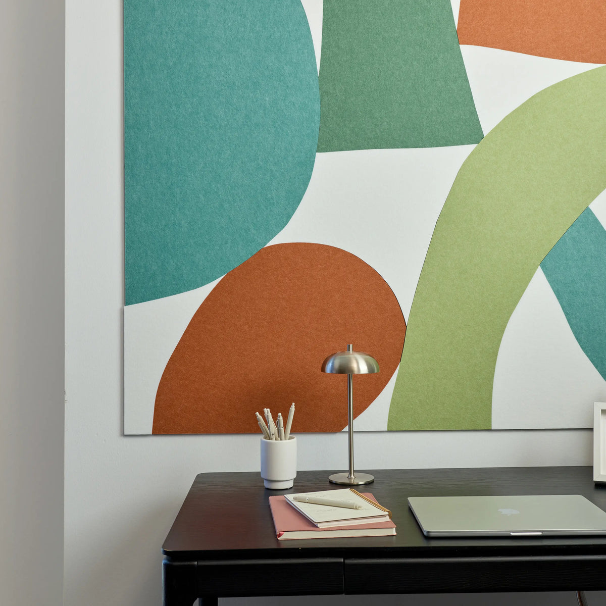 Large abstract acoustic wall art with overlapping organic shapes in teal, green, orange, and light green on a white background, displayed above a dark desk with a lamp, notebook, and closed laptop.