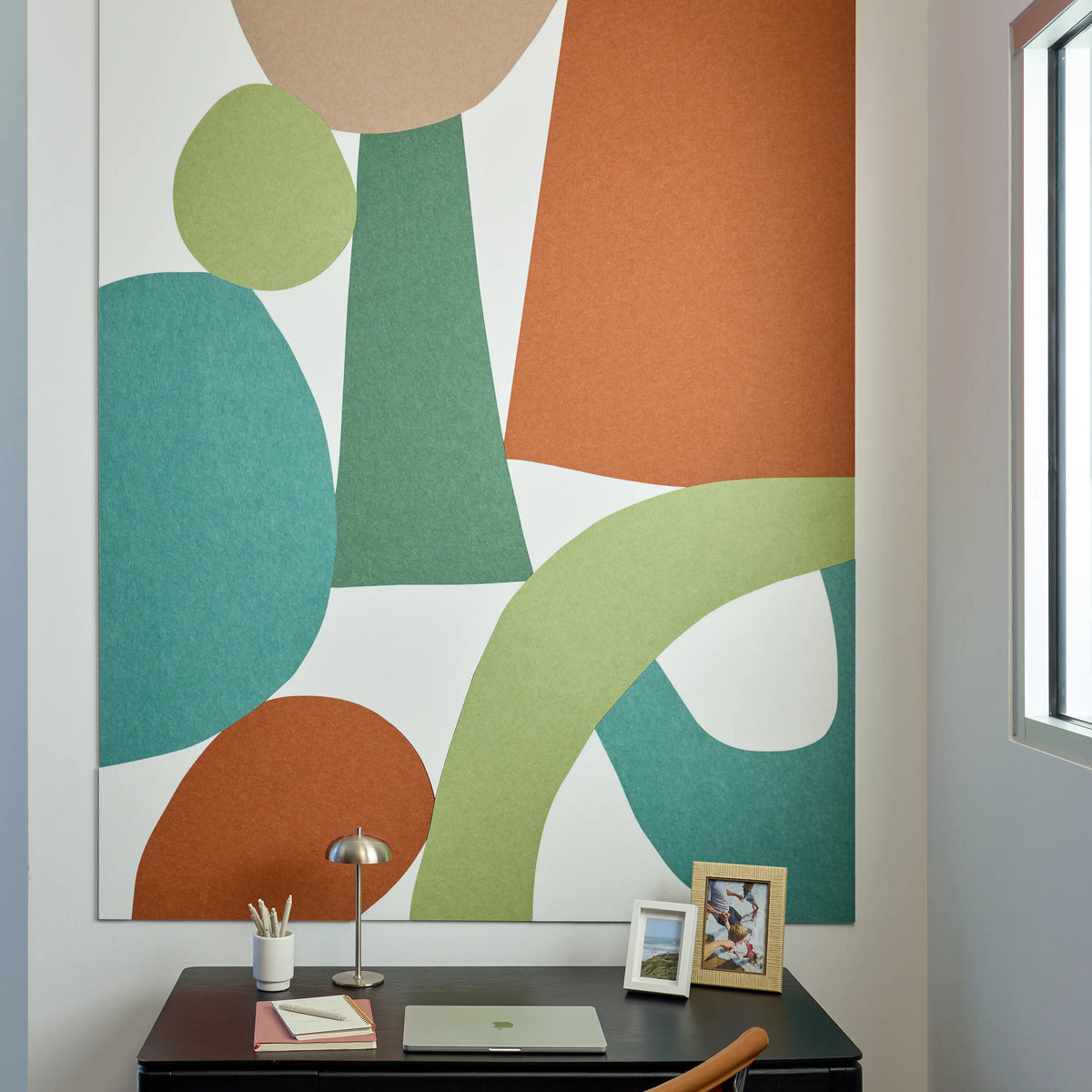 Large abstract wall art with bold organic shapes in teal, sage green, peach, tan, and white, displayed above a dark desk in a bright home office setting.