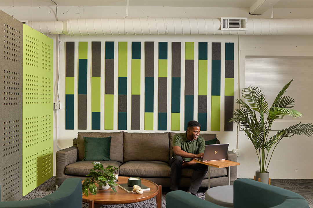 A Felt Right wall art design in shades of grey and green above a couch on the wall in a stripe like pattern and a guy working on his computer