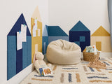 Felt Right wall art in a play room on a white wall in the shape of little houses using shades of blue and yellow felt tiles in a corner of a room with a rug and some kid toys.