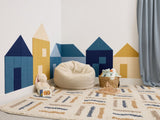 Felt Right wall art in a play room on a white wall in the shape of little houses using shades of blue and yellow felt tiles in a corner of a room with a rug and some kid toys.