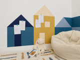 Felt Right wall art in a play room on a white wall in the shape of little houses using shades of blue and yellow felt tiles in a corner of a room with a rug and some kid toys.