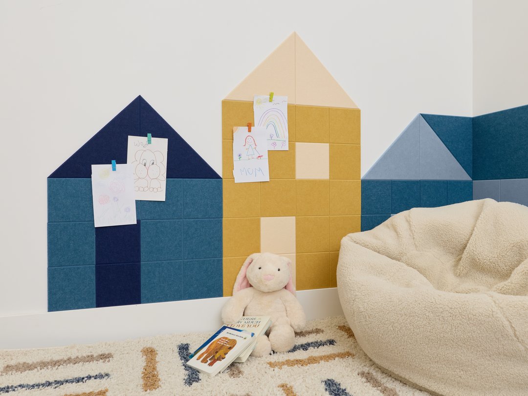 Felt Right wall art in a play room on a white wall in the shape of little houses using shades of blue and yellow felt tiles in a corner of a room with a rug and some kid toys.