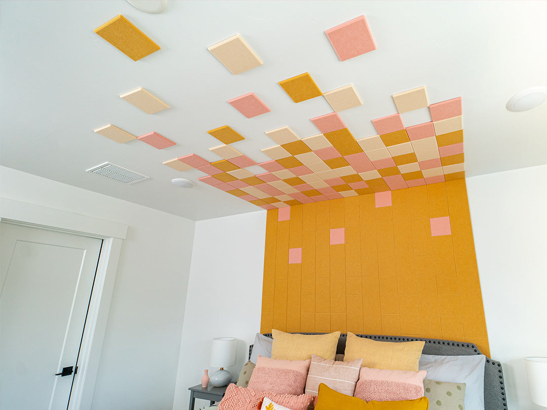 A Felt Right wall art design in shades of yellow, pink, and cream square felt tiles in a bedroom on a white wall and on the ceiling above a bed.