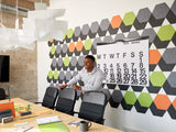 A Felt Right wall art design in shades of grey, orange, and green in a hexagon shapes on a white wall in a conference room or an office with a giant calendar and a man by the chairs