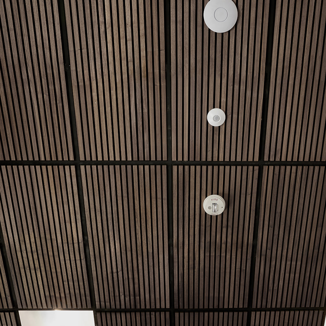 A close-up view of a dark brown slatted ceiling with black grid lines and three white circular smoke detectors mounted vertically.