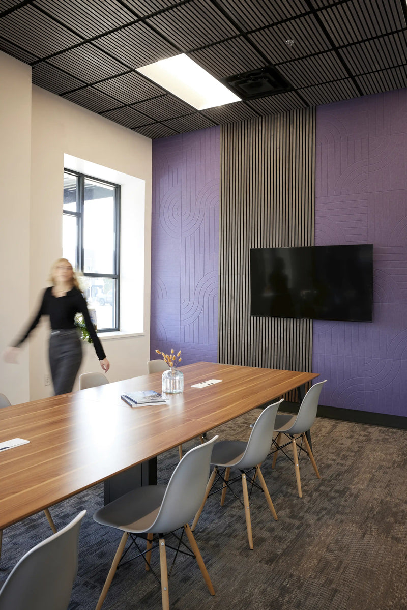 A modern conference room with a long wooden table, gray chairs, a black slatted ceiling, and purple accent walls. A woman is blurred in motion near the window on the left.