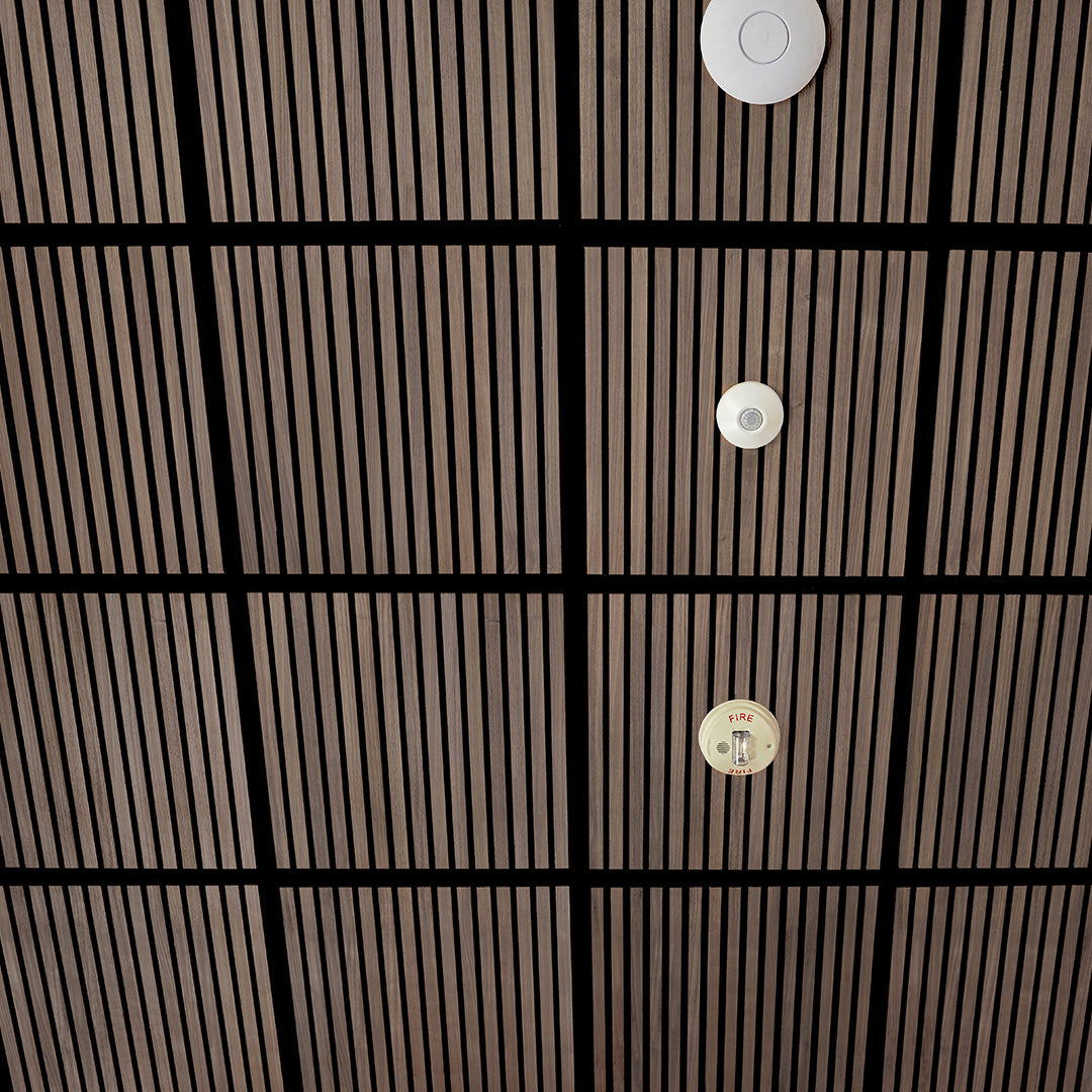 A close-up view of a modern ceiling with a grid of dark wood slats and black beams, featuring three circular white and beige ceiling-mounted fixtures.