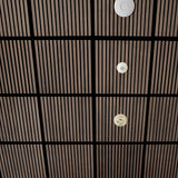 A close-up view of a modern ceiling with a grid of dark wood slats and black beams, featuring three circular white and beige ceiling-mounted fixtures.