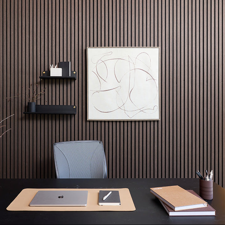A modern home office setup featuring a dark wood slatted accent wall with a minimalist abstract art print hanging above a desk. The desk is styled with a beige desk mat, a closed silver laptop, a small notebook, and stacked notebooks, with a gray office chair in the background.