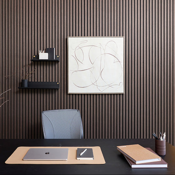 A modern home office setup featuring a dark wood slatted accent wall with a minimalist abstract art print hanging above a desk. The desk is styled with a beige desk mat, a closed silver laptop, a small notebook, and stacked notebooks, with a gray office chair in the background.