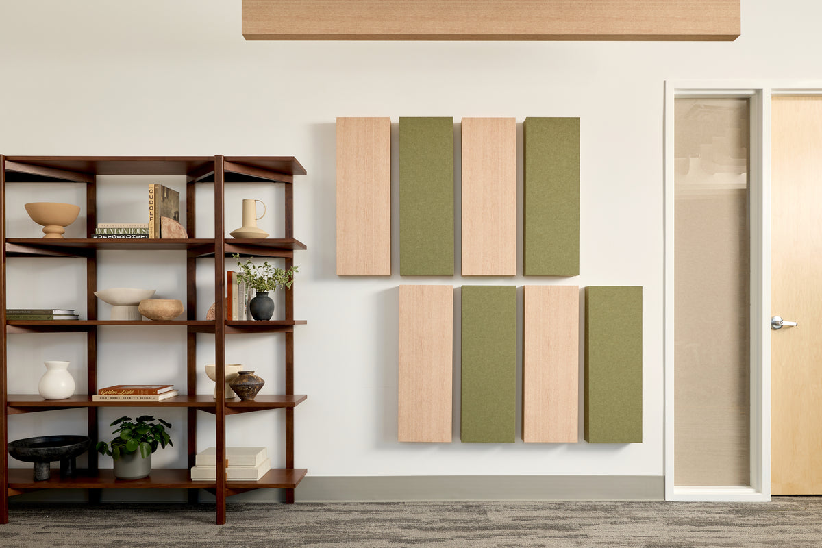Wall-mounted acoustic panels in light wood and sage green arranged on a white wall in a modern office interior, with a wooden shelving unit on the left and a glass door on the right.