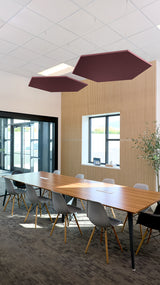 Two burgundy hexagonal acoustic baffles suspended from ceiling in modern office space above wooden table.