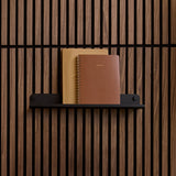 Black wall shelf with rounded ends mounted on vertical wood slat wall, holding two notebooks.