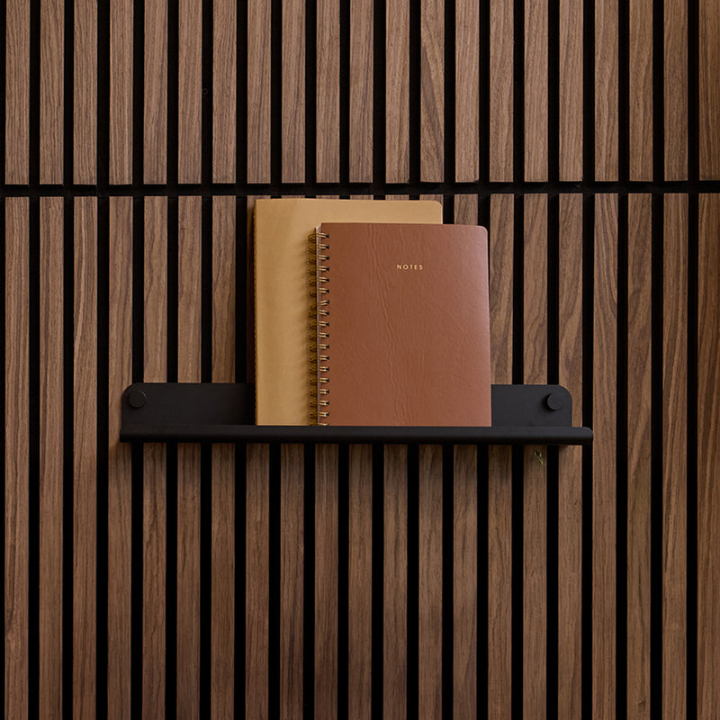 Black wall shelf with rounded ends mounted on vertical wood slat wall, holding two notebooks.